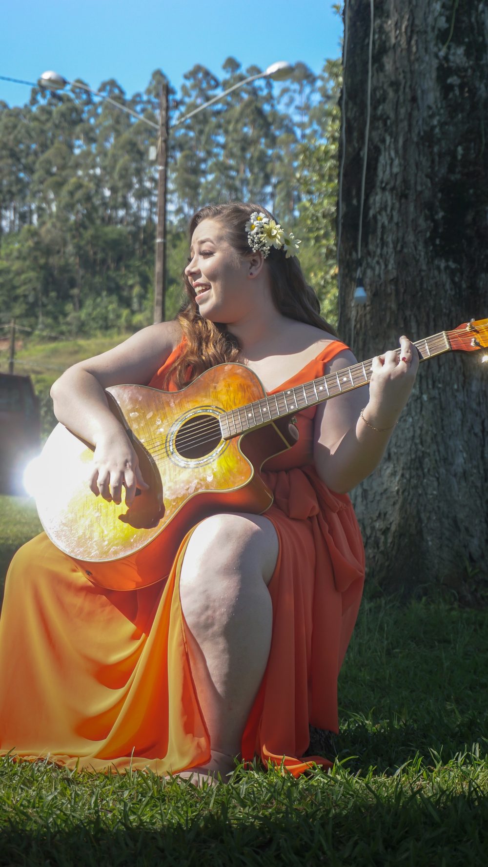 Letícia Bertoldi tocando violão ao ar livre, de vestido laranja